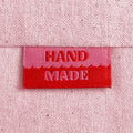 A close-up of a pink and red woven sewing label with the words 'HAND MADE', sewn into light pink canvas.