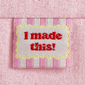A close-up of a pink striped woven sewing label with a scalloped yellow border around red text reading 'I made this!'. The label is sewn into a light pink canvas fabric.