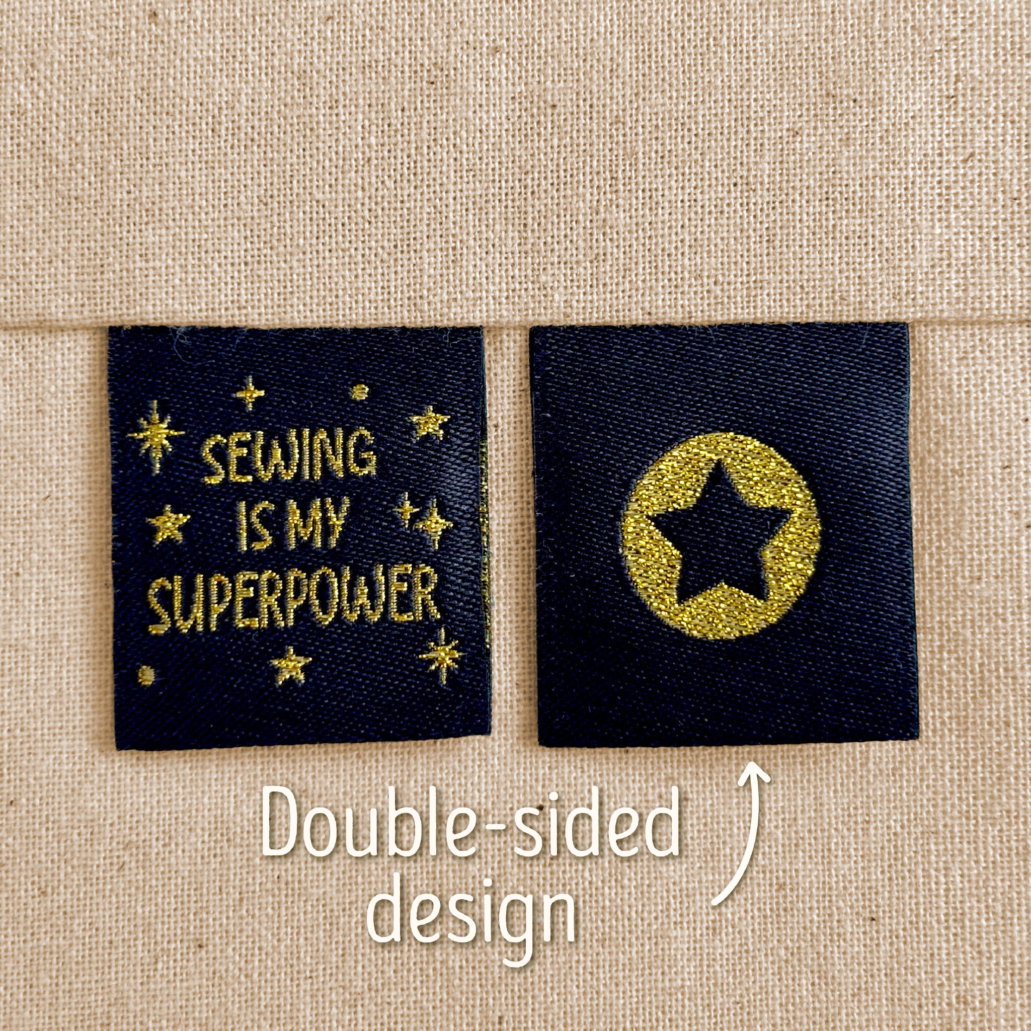Two black woven sewing labels that have been sewn into beige canvas. The left label reads “Sewing Is My Superpower” in sparkly gold lettering, which surround by gold stars. The right label shows a black star inside a sparkly gold circle. Text in the image explains that this is a double-sided design, implying that the star is on the back of the 'Sewing Is My Superpower' design.